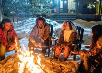 Group of happy friends sitting on chairs and cooking marshmallows near the fire during their winter vacation in the country house