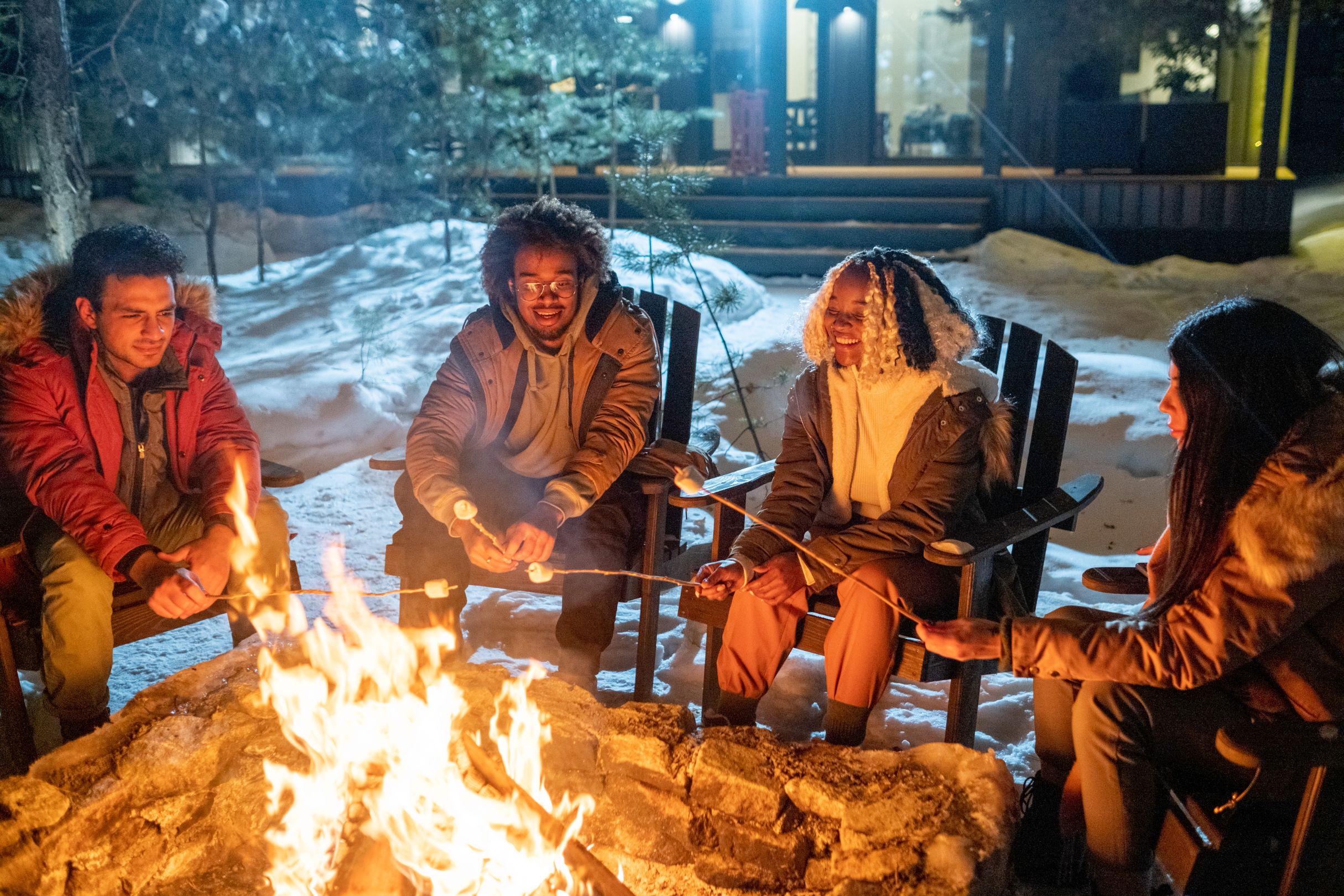 Group of happy friends sitting on chairs and cooking marshmallows near the fire during their winter vacation in the country house
