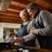 Elderly multi-ethnic couple smiling while cooking together in their modern kitchen. Happy and healthily retired
