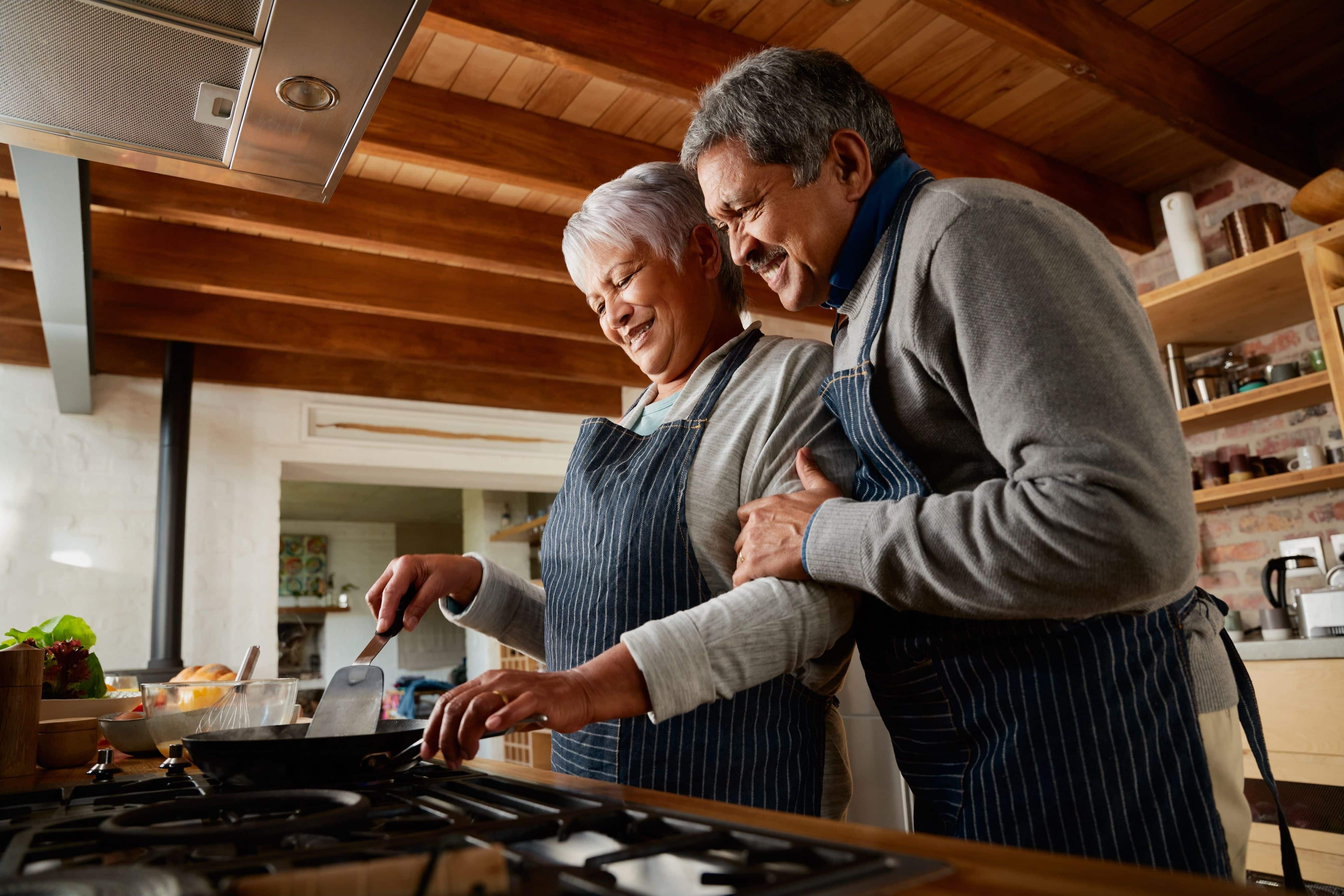 Elderly multi-ethnic couple smiling while cooking together in their modern kitchen. Happy and healthily retired