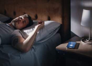 Caucasian Man Sleeps through Smartphone Alarm Clock Showing Eight in the Morning. Tired Person Oversleeping. Person and Mobile Phone Close-up. Bedside Nightstand Bedroom Apartment