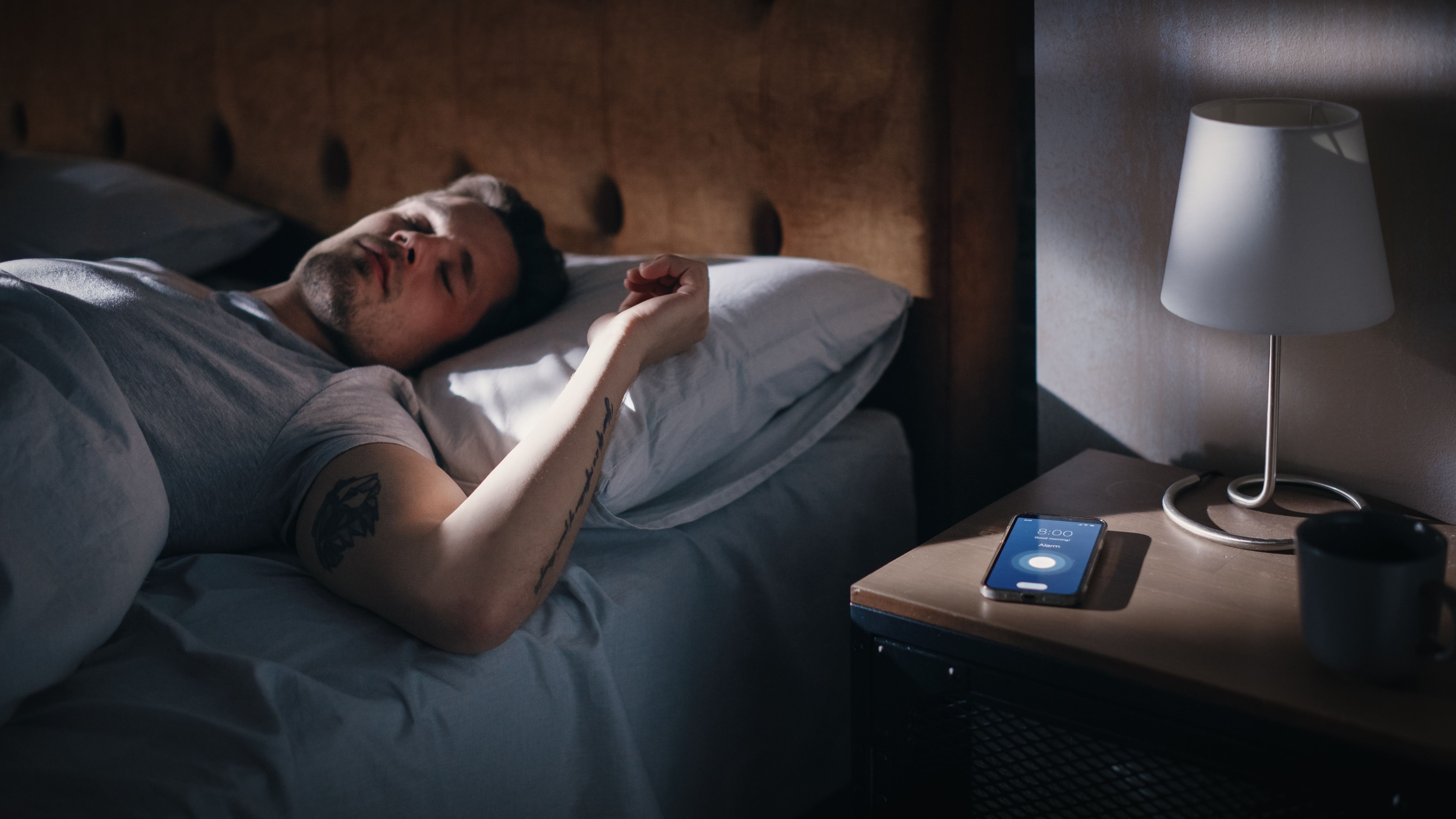 Caucasian Man Sleeps through Smartphone Alarm Clock Showing Eight in the Morning. Tired Person Oversleeping. Person and Mobile Phone Close-up. Bedside Nightstand Bedroom Apartment