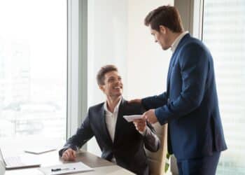 General manager presenting an envelope with premium or bonus cash to male company official. Boss congratulating happy employee with career promotion, thanking for good job and giving financial reward