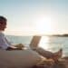 Senior man working on his laptop lying on deck chair on the beach during sunset