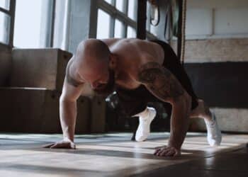 Young strong muscular shirtless Caucasian male athlete doing burpee exercises during functional workout in large gym.