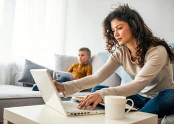 Smiling mom working at home with her child on the sofa while writing an email. Young woman working from home, while in quarantine isolation during the Covid-19 health crisis