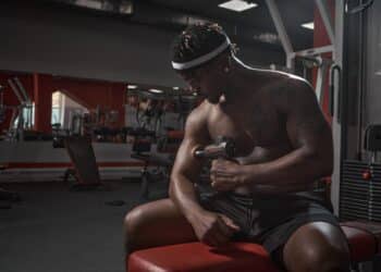 Athletic african american man massaging hand by handheld massage gun, post-workout recovery routines in gym