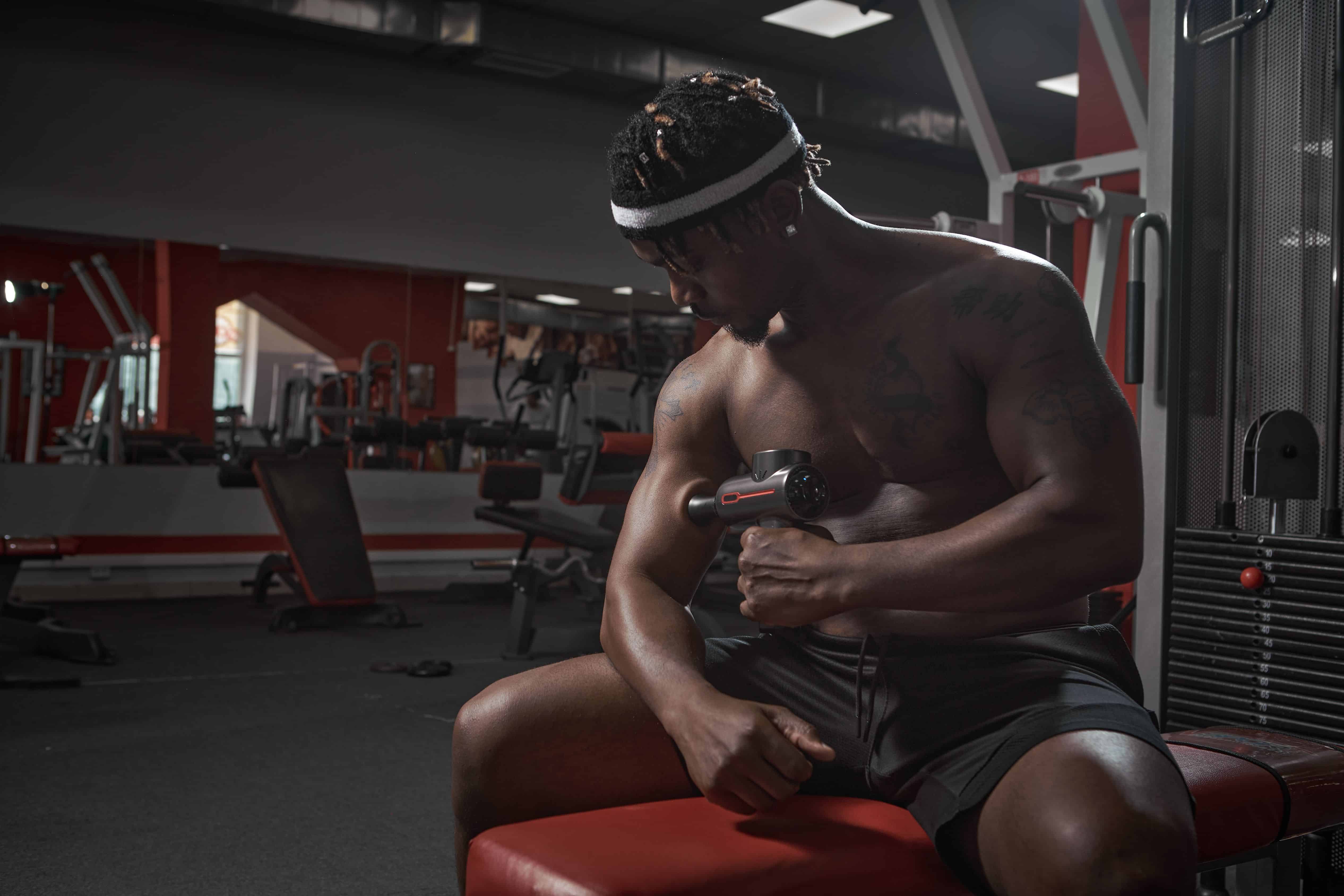 Athletic african american man massaging hand by handheld massage gun, post-workout recovery routines in gym