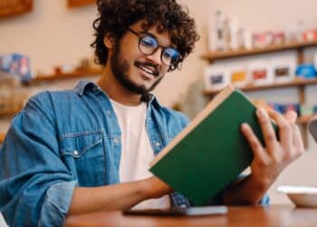 Young indian handsome man in glasses reading book at cafe