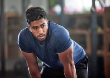 Tired, breathing and fitness gym man taking a break from workout, training or exercising inside a wellness center. Young athletic, masculine guy resting after his strength or muscle exercise session