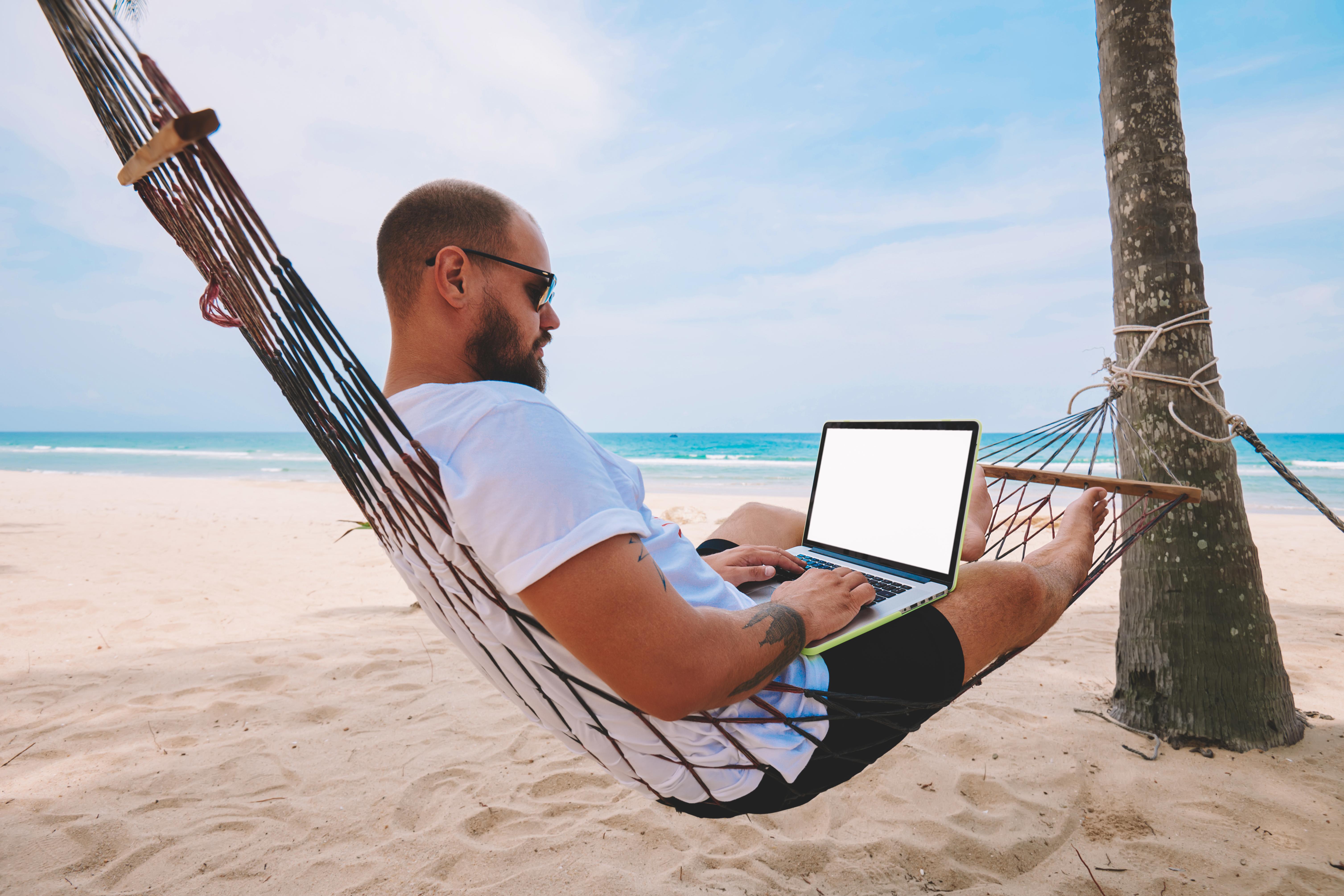 Young bearded businessman in sunglasses rest on hammock on ocean beach while working distantly on laptop computer with blank screen area for your web page advertise content during summer vacations