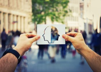 Man hands holding a white paper sheet with two faced head over a crowded street background. Split personality, bipolar mental health disorder concept. Schizophrenia psychiatric disease.