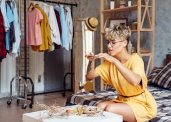 Young woman food blogger wearing eyeglasses sitting at stylish urban apartment taking photo of plate with desserts macaroons and eclairs on smartphone smiling joyful