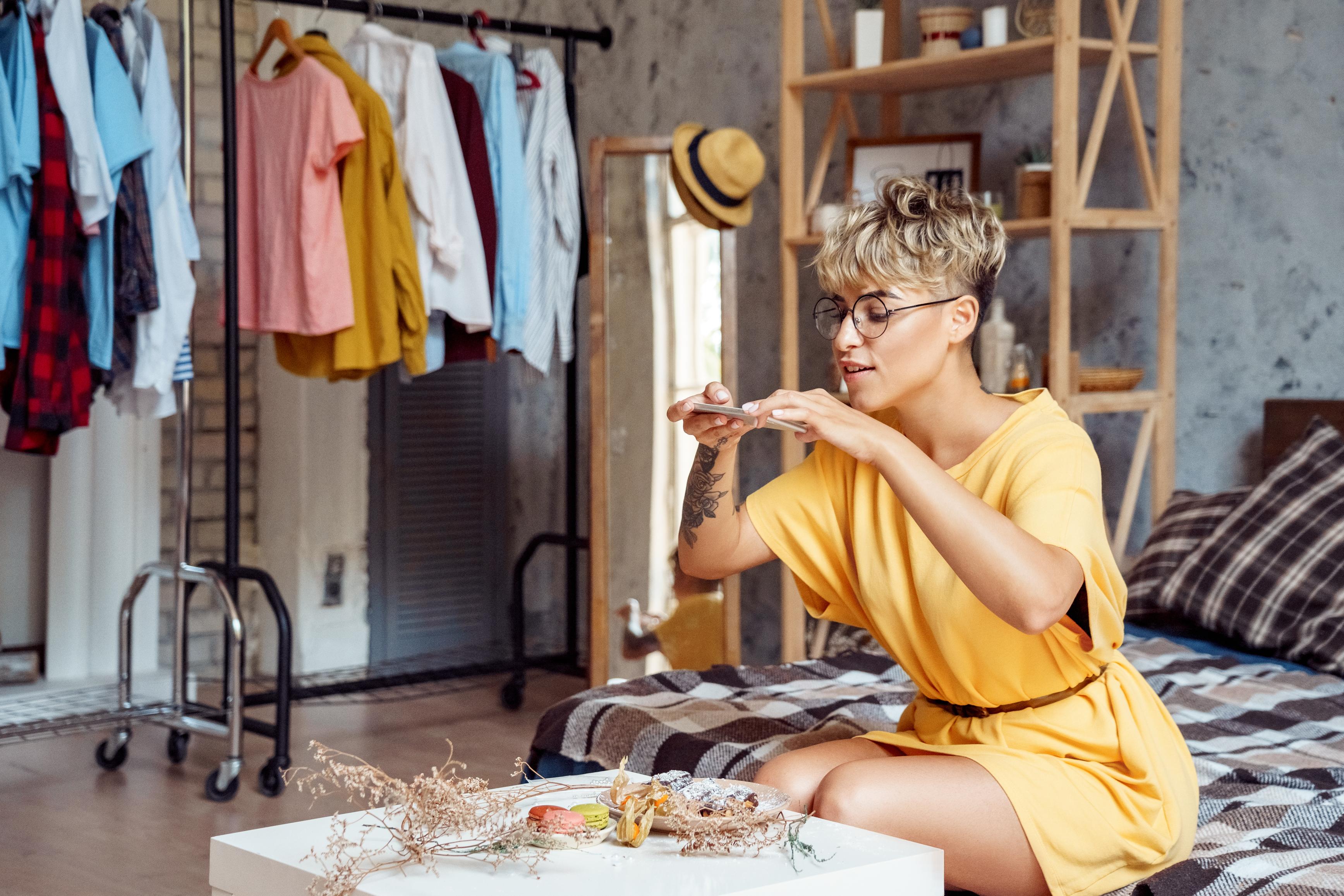 Young woman food blogger wearing eyeglasses sitting at stylish urban apartment taking photo of plate with desserts macaroons and eclairs on smartphone smiling joyful