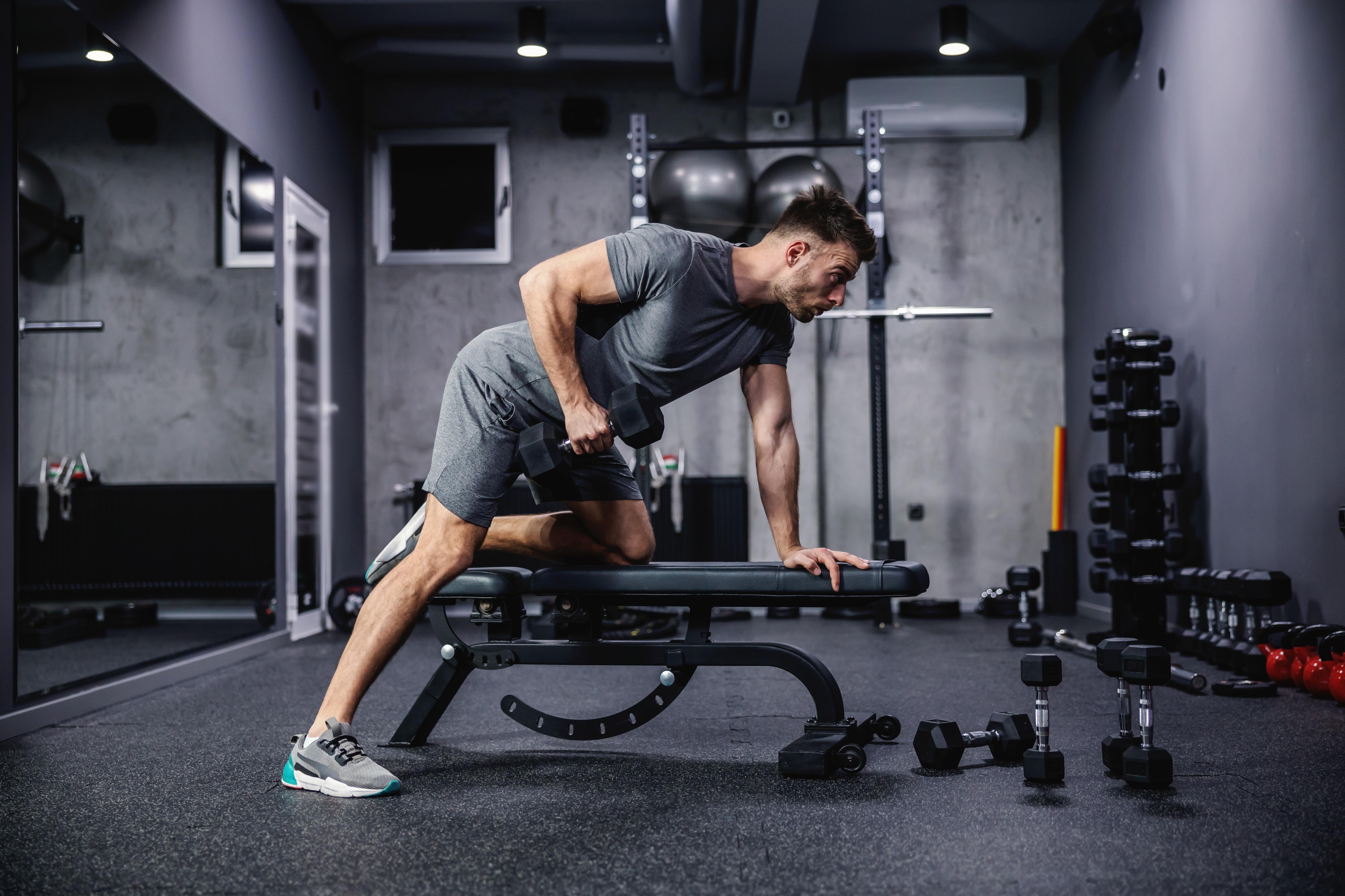 Man doing exercise with dumbbell leaning on sports bench in the gym. Photo of a sexy muscular man in sportswear and good physique on grey background. Strength and motivation, sport, fitness goal
