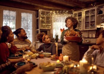 Happy African American mature woman brining stuffed turkey at dining table during family Thanksgiving dinner.