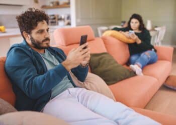 Domestic Tension: Man Engrossed in Cellphone with Disapproving Wife - Man on cellphone; wife's disapproving look captures modern relationship tension.