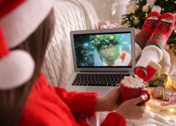 MYKOLAIV, UKRAINE - DECEMBER 25, 2020: Woman with sweet drink watching The Grinch movie on laptop at home, closeup. Cozy winter holidays atmosphere
